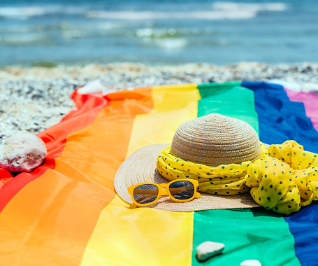a straw hat with a yellow scarf and sunglasses, over a rainbow flag and the sea in the background. summer vacations in the mediterranean sea. lgtb concept.