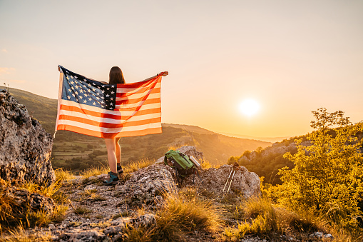 Young woman waving a USA flag at the top of the mountain at sunset.