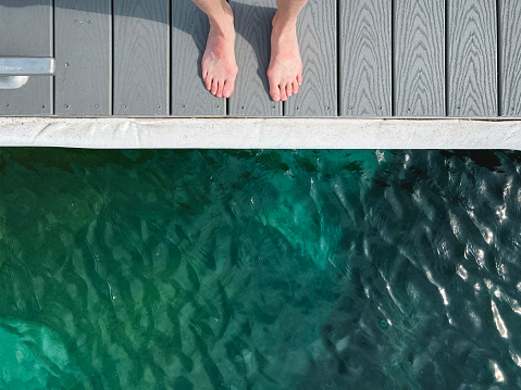 Looking down over a turquoise body of crystal clear waters. A pair of human feet stand at the edge of a dock or pier. Person needs to decide whether to jump in or remain standing on the dock. Concept for the decisions of life - whether to thrive or remain stagnant.