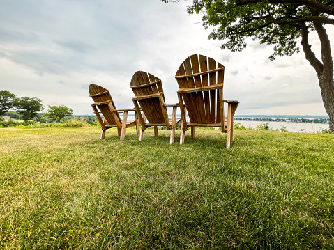 Rear view of three wooden high-back Adirondack chairs resting on the green summer grass. Chairs are situated on a bluff overlooking a harbor on Lake Michigan. Located in the summer getaway of Harbor Springs, Michigan, USA.