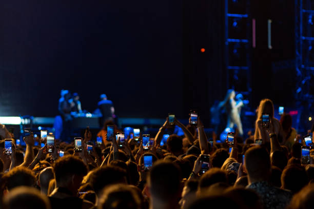 vista posteriore ravvicinata di una grande folla di persone che si godono un concerto all'aperto in una notte d'estate. ci sono molte mani alzate che tengono gli smartphone e registrano lo spettacolo invece di guardarlo effettivamente. - youth culture city life urban scene teenager foto e immagini stock