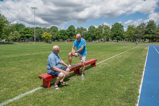 Two old men are having a conversation outdoors on a sports field. One man is sitting whilst the other is standing with one leg up on the bench. This is a married, gay couple.