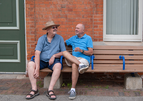 Two men, a senior citizen LGBT married gay couple sit on an outdoor train station bench with a red brick wall behind them.