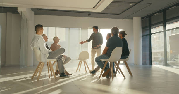 hombres de negocios sentados en círculo en una reunión de terapia de apoyo con el líder o entrenador dando un discurso de bienvenida. joven o facilitador de pie y participando en una discusión grupal interactiva - mediación fotografías e imágenes de stock