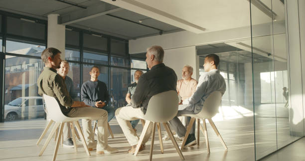 A support group of men talking and sitting in a circle during a therapy session in a workshop or corporate office. Team of colleagues discussing feelings and emotions in a group psychology meeting stock photo