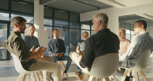 istock Young man in rehab discussing his work and personal problems in a therapy support group session for business workers. Group of businessmen sitting in a circle at a team building meeting 1411325467