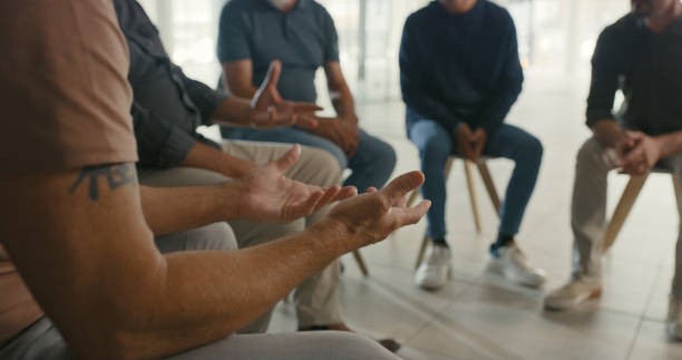 uomo in una riunione di terapia di gruppo che gesticola o chiede aiuto per la sua salute mentale. comunità di uomini si riuniscono e ascoltano il terapeuta discutere i meccanismi di coping in una sessione di supporto sociale - addiction foto e immagini stock