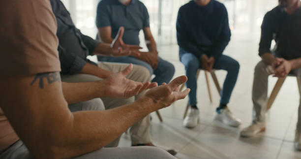 Man in a group therapy meeting gesturing or asking for help with his mental health. Community of men gather round and listen to therapist discuss coping mechanisms in an social support session stock photo