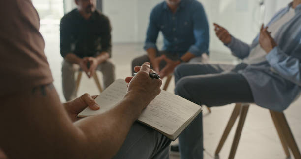 istock Psychology therapist writing notes in book for group therapy with men. Listening to shared trauma experiences and stories with diverse people. Supported in routine meeting for mental help from peers 1411325493