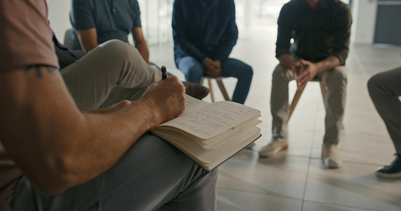 Closeup of a professional psychologist writing notes in a book in during a therapy session. Male therapists analyze patients and write in a notebook while sitting in a circle discussing mental health