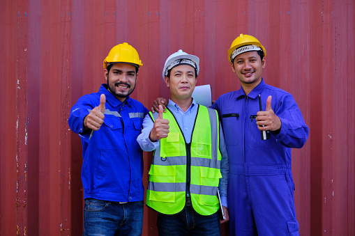 Portrait of three Asian industry workers with safety uniform and hard hats on metal sheet background, thumb up, look at camera and smile, cargo shipping, professional freight transportation business.