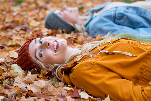 Beautiful young woman laying on the grass outdoors covered in colorful Autumn leaves with her friend. She is looking at the camera and smiling a toothy, white smile and is wearing a knitted hat and a fall jacket.