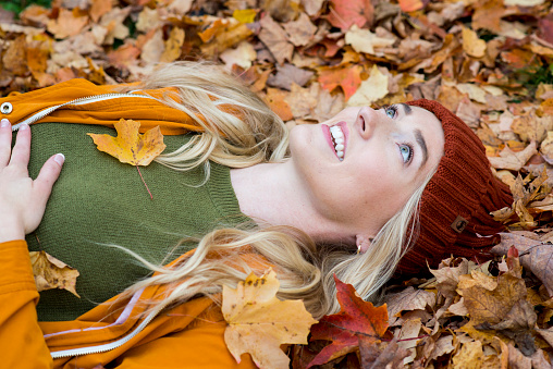 Beautiful young woman wearing an orange fall jacket and green knitted sweater is laying in a pile of fallen, colorful Autumn leaves and looking up at the sky while smiling.
