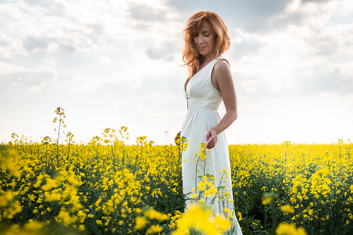 Woman in a long yellow dress walking on the field with yellow flowers at sunset. Summer landscape