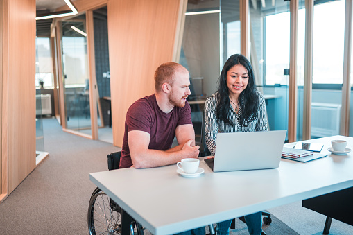 Indoor shot of white man in a wheelchair next to Latin woman seated at a desk. they are using a laptop to write important reports due at night.