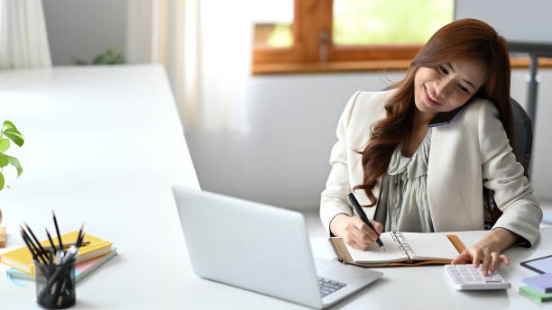 istock Asian working woman having phone conversation and using calculator at her office desk. 1411362587