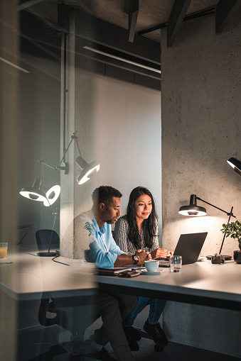 Candid shot of African American male and Hispanic female working hard at the office during the night. They are seated in a desk and their glass door is open reflecting the light from their desk lamp.