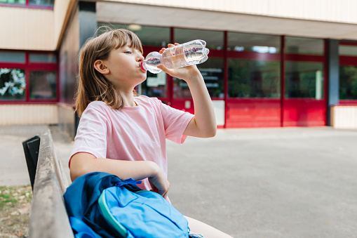 A schoolgirl sits in the school yard and drinks fresh water from a bottle. Water balance of children during school hours. Back to school concept.
