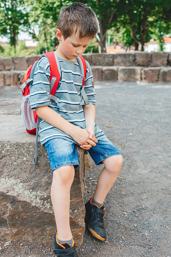 A sad schoolboy sits in the school yard with a backpack on his back. Children's problems at school. The boy does not want to go to school.