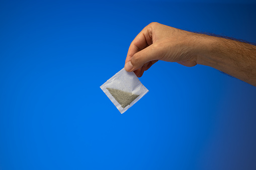 Single white paper tea bag held in hand by Caucasian male hand. Close up studio shot, isolated on blue background.