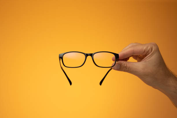 istock Black plastic frame eyeglasses held in hand by Caucasian male hand. Close up studio shot, isolated on orange background 1411368144