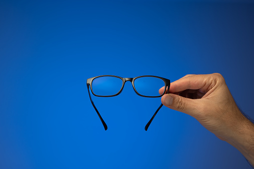 Black plastic frame eyeglasses held in hand by Caucasian male hand. Close up studio shot, isolated on blue background.
