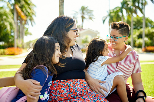 Front view of smiling mid adult women with 5 and 8 year old daughters sitting together on park bench in sunny South Florida.