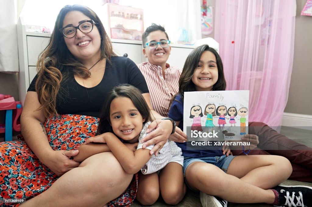 Portrait of lesbian parented family and child’s drawing Front view of early 30s same-sex couple sitting on floor with young girls, all casually dressed and smiling at camera. LGBTQIA Rights Stock Photo