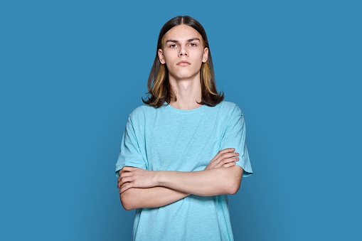 Portrait of confident serious teenage guy looking at camera on blue color background. Handsome young male with crossed arms posing in studio. Youth, students, people concept