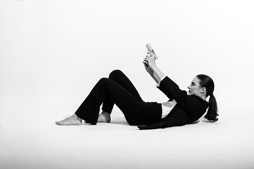 Beautiful woman lying on white studio ground with black classic suit, white shirt and ponytail hairstyle with silver pistol in her hands and pointing it up. Model with bare feet. Black and white image