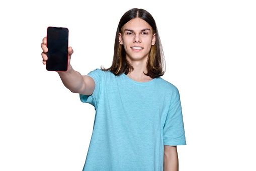 Teenage handsome guy showing blank smartphone screen, on white isolated background. Teen student smiling posing in studio, blank screen space for text, logo, advertisement