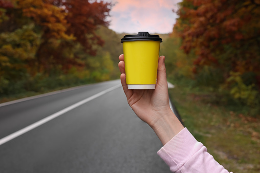 Woman holding takeaway cardboard coffee cup near road outdoors, closeup. Autumn season
