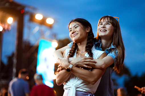 Two girlfriends of Asian and Caucasian ethnicity are hugging and enjoying the atmosphere at a music festival as the night is falling.