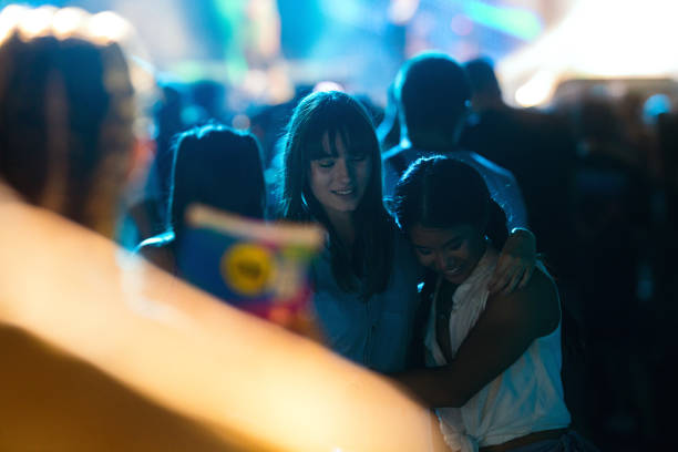 Two girlfriends of Asian and Caucasian ethnicity are hugging and enjoying the atmosphere at a music festival as the night is falling.