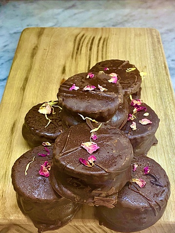 Vertical close up of wood cutting board piled with handmade raw chocolate cherry and coconut bites with Rosewater and edible rose petal garnish