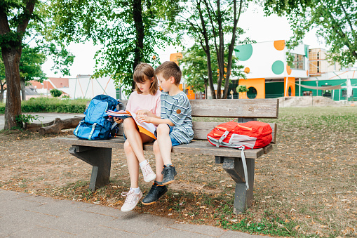 Back to school. Schoolchildren with backpacks sit on a bench in the school yard at recess and deal with homework