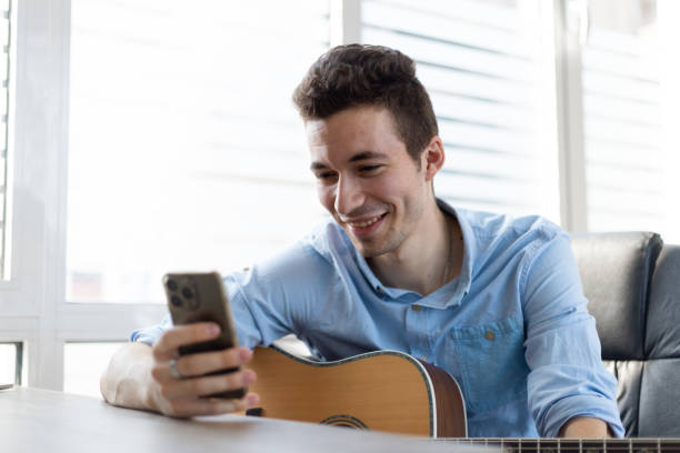 istock Male guitar player sit indoor with smart phone, take break resting from playing string instrument using cellphone smiles read pleasant message. Hobby, pastime, modern wireless technology user concept 1411399104