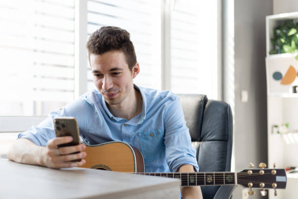 istock Male guitar player sit indoor with smart phone, take break resting from playing string instrument using cellphone smiles read pleasant message. Hobby, pastime, modern wireless technology user concept 1411399119