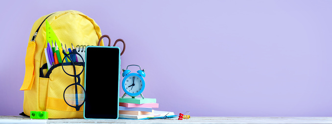 Banner. Concept back to school. Full yellow School Backpack with stationery and smartphone, blue alarm on table on purple background. Panorama.