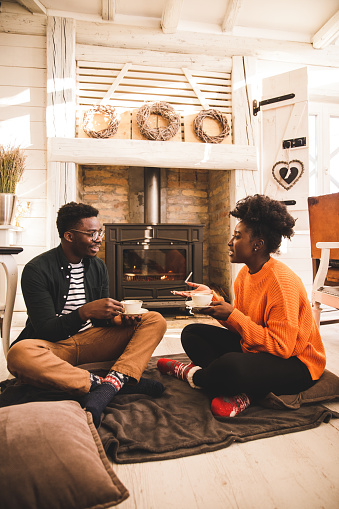 Full length shot of happy young couple sitting on the floor, in a rustic cabin, by the cozy fireplace, enjoying cups of tea and engaging in conversation.