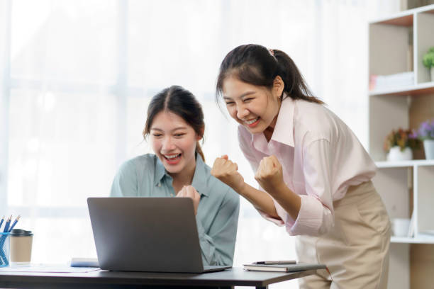 Happy two young Asian businesswomen successful excited raised hands rejoicing with laptop computer in office. New startup project concept. stock photo