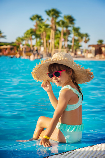 Little girl in a swimsuit, sitting on the edge of the pool, looking at her mobile phone. Childhood, children, smartphone, technology, vacation, internet and fun concept.