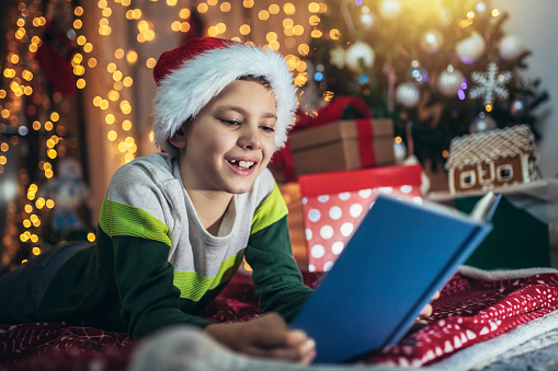 Happy boy lying on front on floor by christmas tree, reading book surrounded by gifts.