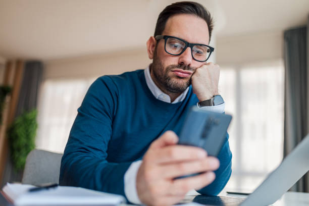 Bored businessman using smart phone while working at the office desk. Bored distracted businessman networking on smart phone while working at desk in the corporate office. boredom stock pictures, royalty-free photos & images