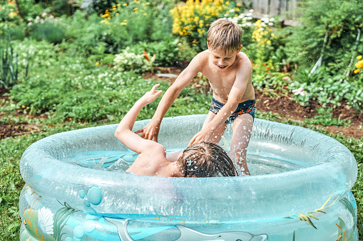 Brothers wrestle in children inflatable pool in cottage yard against lush greenery and flowers. Cheerful schoolboys have fun in water on summer in countryside. No logo