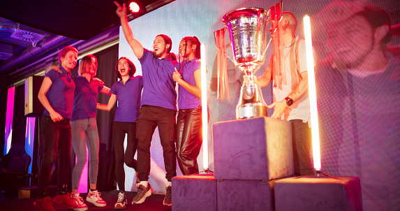 Diverse gamer group wearing blue t-shirts with african ethnicity players and women on stage receiving a trophy. Doing a group cheer and celebrating victory together