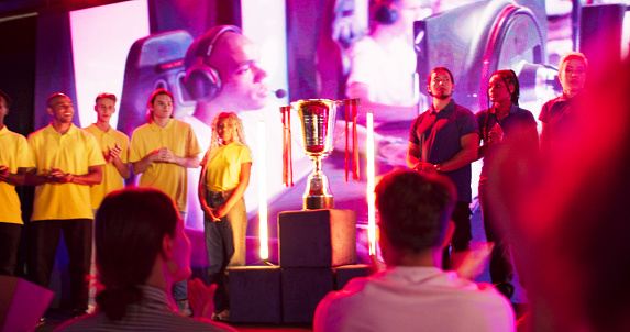 Yellow and blue team standing on a stage and high-fiving. Audience cheering for their favorite players. Trophy standing on the stage.