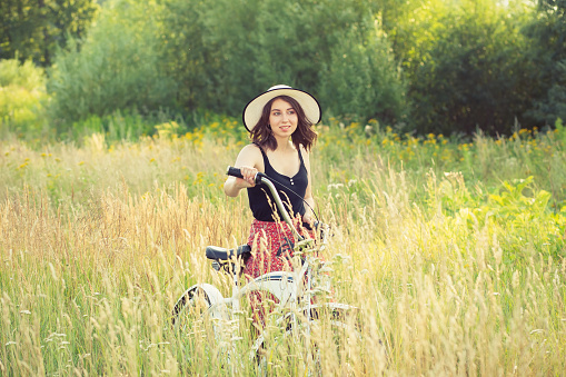 happy brunette girl in hat walking with bicycle at sunny meadow, looking at camera