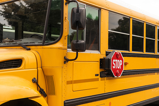 Car point of view looking through the windshield at the rear of a stopped yellow school bus with its automated stop signs and caution lights flashing while students enter or exit the vehicle. It's a rainy, snowy February day near Rochester, New York.