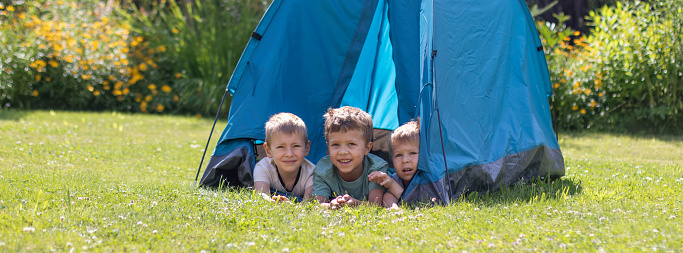 Three little boys lie in a blue tourist tent in the yard. The concept of sports, summer holidays, hiking and happy childhood.